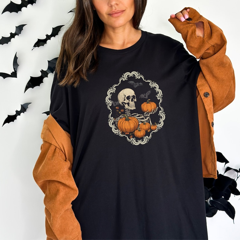 Op de afbeelding: Zwart T-shirt met een afbeelding van een skelet dat pompoenen vasthoudt. De afbeelding is omgeven door een decoratief frame met vleermuizen en bladeren.