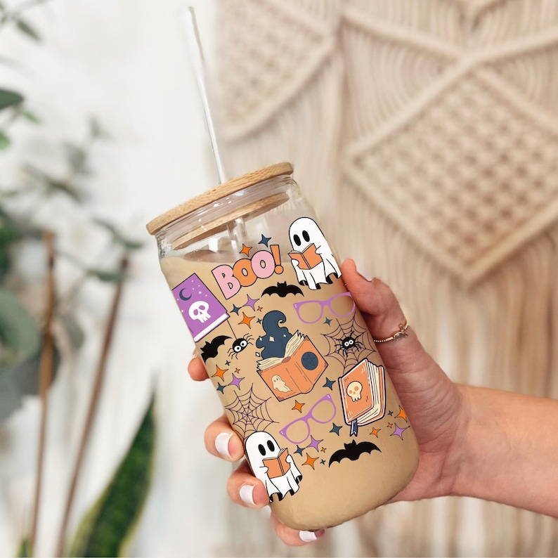 Puede incluir: Un vaso de cristal transparente con tapa de bamb&uacute; y pajita de cristal, lleno de una bebida color canela. El exterior presenta un estampado colorido de Halloween con fantasmas leyendo, ara&ntilde;as, telara&ntilde;as, murci&eacute;lagos, gafas moradas y "Boo!".
