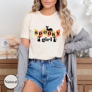 May include: A white t-shirt with a retro-style graphic featuring the text "SPOOKY girl" and black cat silhouettes. The graphic is in shades of orange, yellow, and black.