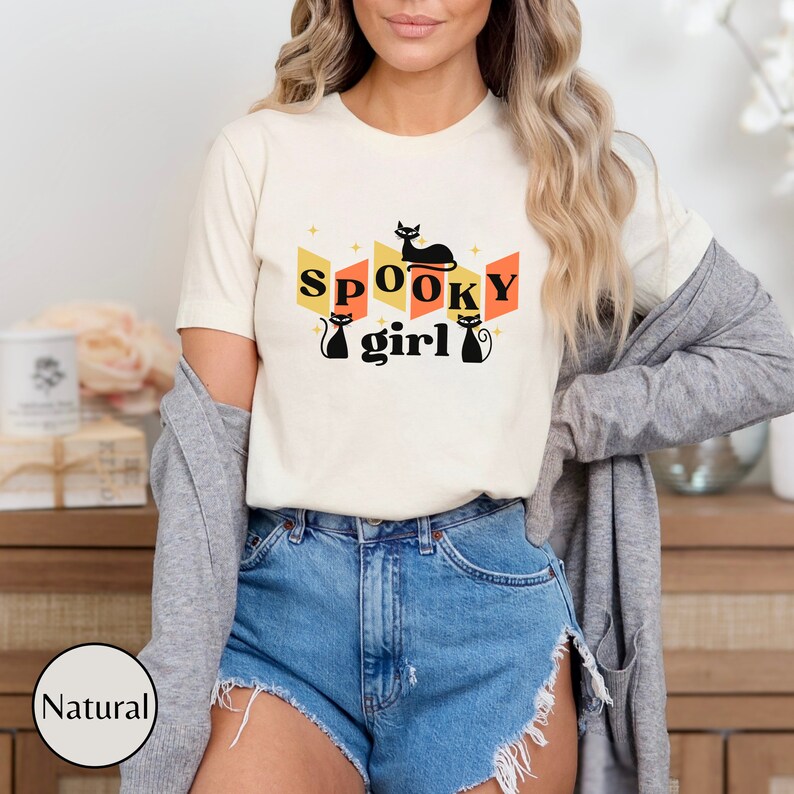 May include: A white t-shirt with a retro-style graphic featuring the text "SPOOKY girl" and black cat silhouettes. The graphic is in shades of orange, yellow, and black.