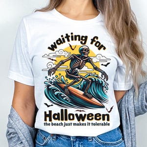 May include: White t-shirt with a Halloween-themed graphic. The design features a skeleton surfing on a wave, with the words "waiting for Halloween" and "the beach just makes it tolerable". The artwork includes bats, a sun, and ocean waves.