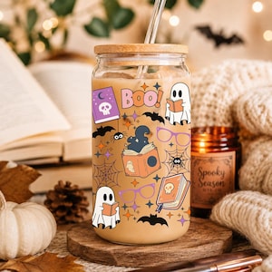 Puede incluir: Un vaso transparente con tapa de bamb&uacute; y pajita de cristal, lleno de una bebida color canela. El exterior presenta un estampado colorido de Halloween con fantasmas, libros, ara&ntilde;as, telara&ntilde;as, murci&eacute;lagos, gafas moradas y el texto BOO en letras rosas.