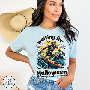 May include: Ice blue t-shirt with a surfing skeleton graphic. The design features a skeleton riding a surfboard on a wave, with the words "waiting for Halloween" and "the beach just makes it tolerable."