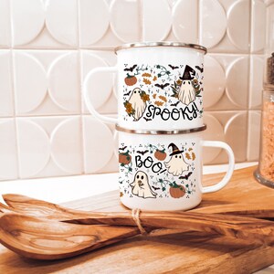 May include: Two white enamel mugs with a Halloween theme. The top mug has a ghost wearing a witch hat and the text "Spooky!" The bottom mug has a ghost holding a flower and the text "Boo!" Both mugs feature a pattern of ghosts, bats, pumpkins, and leaves.