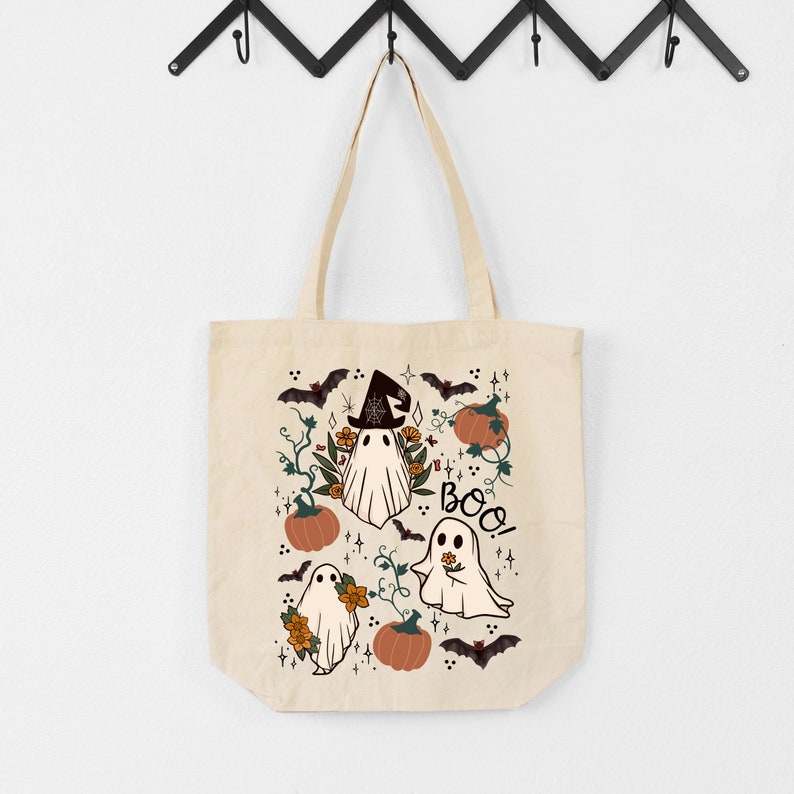 K&ouml;nnte beinhalten: Beige Tragetasche mit einem Halloween-Motiv. Das Design umfasst Cartoon-Geister, K&uuml;rbisse, Flederm&auml;use und florale Elemente. Das Wort "Boo!" ist in einer verspielten Schriftart geschrieben. Die Tasche hat eine einfache, rechteckige Form mit zwei Griffen.