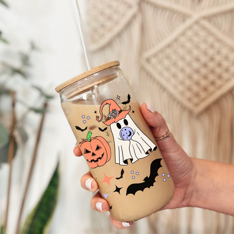 May include: A clear glass tumbler with a wooden lid and a straw. The tumbler is filled with a brown liquid and has a Halloween-themed design featuring a ghost, a pumpkin, and bats.