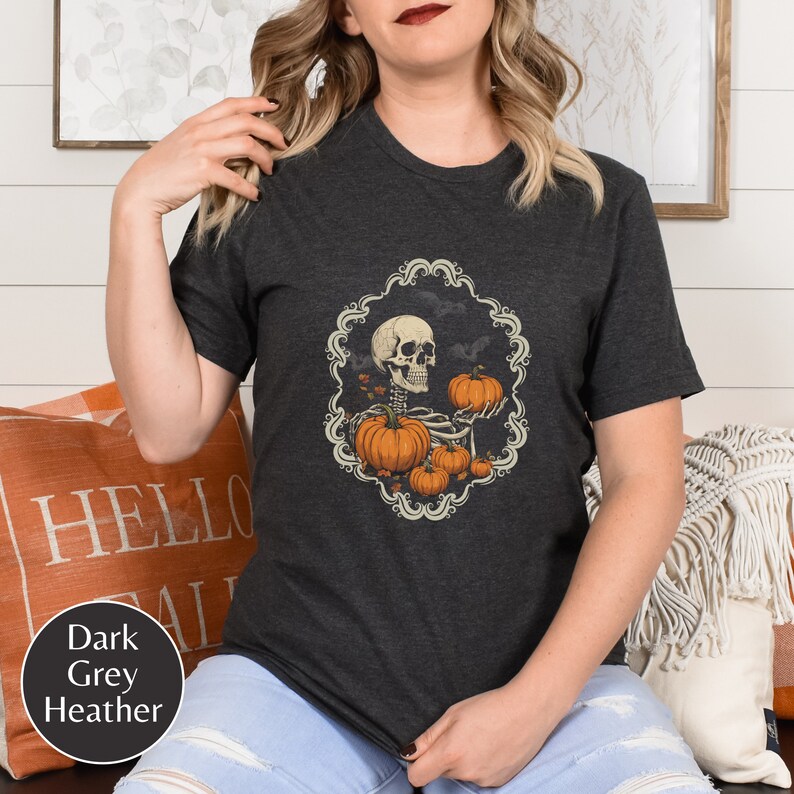 Op de afbeelding: Een donkergrijs heather t-shirt met een afbeelding van een skelet dat een pompoen vasthoudt. Het skelet is omringd door pompoenen en bladeren in een decoratief kader.