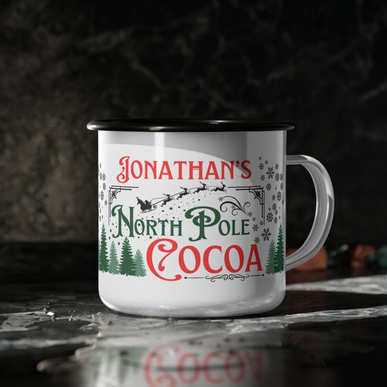 May include: White enamel mug with a black rim and handle. The mug has a Christmas-themed design with the text "Jonathan's North Pole Cocoa" in red, green, and black lettering. The design also includes a silhouette of Santa Claus flying in a sleigh with reindeer.