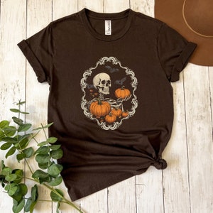 Op de afbeelding: Een bruin T-shirt met een skelet dat pompoenen vasthoudt in een decoratief frame. Het frame is versierd met vleermuizen en bladeren rond het skelet. Dit T-shirt is perfect voor Halloween.