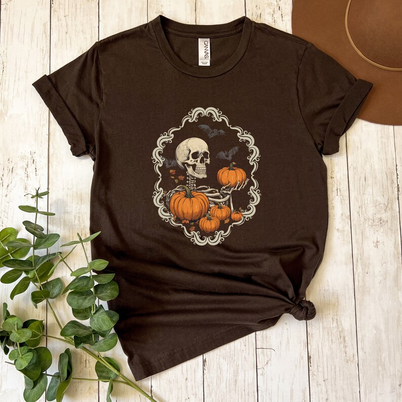 Op de afbeelding: Een bruin T-shirt met een skelet dat pompoenen vasthoudt in een decoratief frame. Het frame is versierd met vleermuizen en bladeren rond het skelet. Dit T-shirt is perfect voor Halloween.