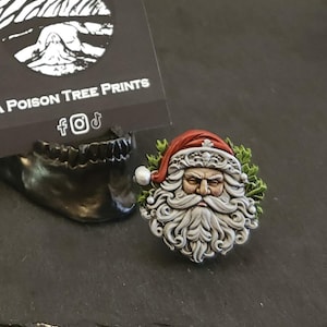 May include: A detailed Santa Claus face pin with a red hat, white beard, and green wreath. The pin is set against a dark background. A business card with the text "A Poison Tree Prints" is in the background.
