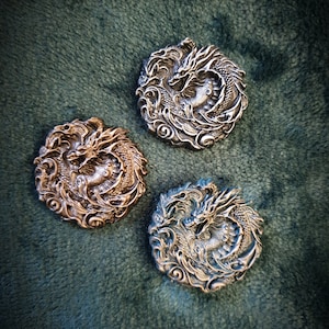 May include: Three round, silver-toned metal ornaments featuring a detailed dragon design. The dragons are curled up in a circle with their tails in their mouths. The ornaments are displayed on a green, fluffy surface.