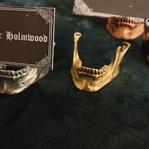 May include: Three metal skull jaw bone card holders. The card holders are in silver, gold, and bronze. The silver card holder has a black card with the text "Arthur Holmwood" on it.