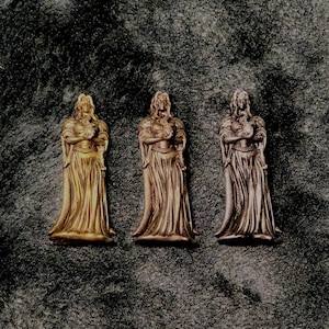 Puede incluir: Tres estatuas metálicas de una figura vestida con una túnica y pelo largo, cada una en un tono diferente de oro, bronce y cobre. Las estatuas están de pie, con las manos juntas frente a ellas. El fondo es una superficie oscura y texturizada.