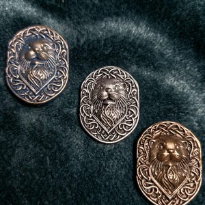 May include: Three oval metal buttons featuring otter faces within Celtic knotwork borders. The buttons are in bronze, silver, and blue hues, set against a dark green velvet backdrop. The otter's face is intricately detailed.