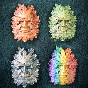 May include: Four Green Man masks made of resin. The masks are all different colours: orange, green, bronze, and rainbow. Each mask has a face with a beard and is surrounded by leaves.