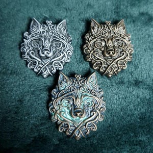 以下が含まれることがあります： 複雑なケルトノットのデザインが施された、金属製のオオカミの頭のペンダント3つ。1つはシルバー、1つはブロンズ、3つ目はターコイズとブロンズの仕上げです。ペンダントは濃い緑色のテクスチャのある表面に表示されています。