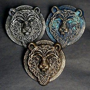 May include: Three circular bear head medallions with detailed Celtic knotwork. The medallions are in silver, blue, and bronze hues. The bear heads have a raised relief design, showcasing a blend of artistry and craftsmanship. The overall design is intricate and detailed.