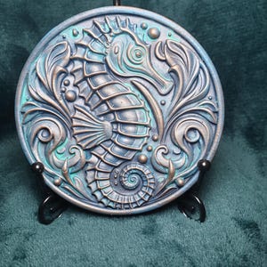 May include: A circular, blue and bronze decorative item with a seahorse motif. The seahorse is centrally positioned, encircled by swirling, stylised aquatic plants. It is displayed on a black metal stand.