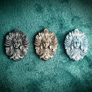 May include: Three decorative wall plaques featuring a stylised female face with a headdress. The plaques are made of metal and are painted in silver, gold, and blue.