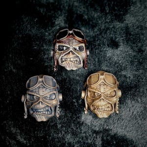 Peut inclure: Trois figurines de crânes métalliques avec des lunettes d'aviateur. Les crânes ont un aspect vieilli, avec des détails comme les dents et les rides. L'un est en bronze, l'autre en argent et le troisième est un mélange des deux.