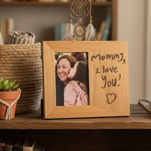 May include: A wooden picture frame with a photo of a woman being kissed by a child. The frame has the handwritten text "Mommy, I love You!" and a heart symbol. The frame is on a wooden shelf with other decorative items.