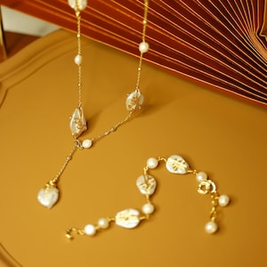 May include: A gold necklace and bracelet set with freshwater pearls and irregular, off-white pendants. The jewellery is displayed on a gold tray, with a decorative fan in the background.
