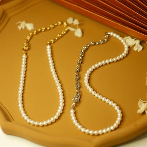 May include: Two pearl necklaces displayed on a brown tray. One necklace has a gold chain, and the other has a silver chain. The pearls are white and round. The background includes a decorative fan and dried flowers.