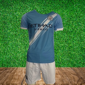 May include: A light blue soccer jersey with white accents and the text "ETIHAD AIRWAYS" on the front. The jersey has the Manchester City crest on the left chest. The jersey is paired with white soccer shorts.