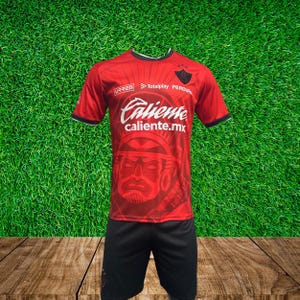 May include: A red soccer jersey with black trim and a black and white graphic of a warrior's face. The jersey has the text "Caliente caliente.mx" and the logos of "Totalplay" and "Urrea".