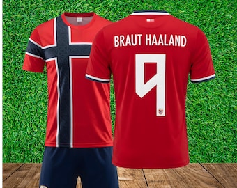 May include: A red and navy blue soccer jersey set featuring the Norwegian flag pattern on the front. The back of the jersey displays the name BRAUT HAALAND and the number 9 in white. The navy blue shorts include a small Norwegian flag emblem.
