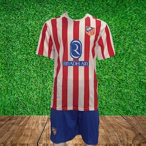 May include: A soccer uniform set featuring a red and white striped jersey with the "Riyadh Air" logo and a team emblem. The shorts are blue with a team logo. The uniform is displayed on a wooden surface against a green background.