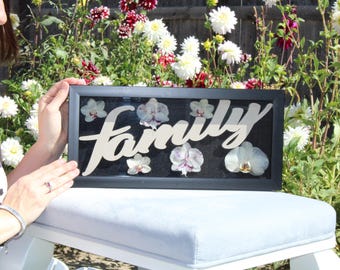 Orquídeas blancas y moradas preservadas con letrero familiar de madera en una vitrina negra.