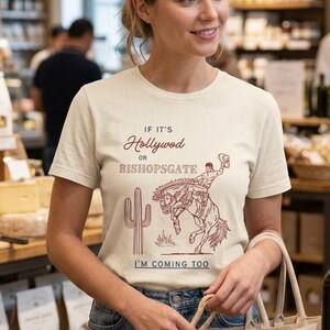 May include: A woman wearing a cream-colored t-shirt with a maroon graphic of a cowboy on a bucking bronco and the text "If it's Hollywood or Bishopsgate, I'm coming too." She is holding a shopping bag in a store.