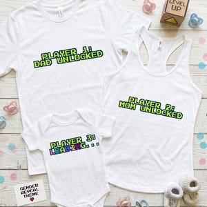 May include: White t-shirts and a onesie with pixelated text. The adult shirts read "Player 1: Dad Unlocked" and "Player 2: Mom Unlocked." The onesie says "Player 3: Loading..." A "Gender Reveal Theme" card and baby items are also present.
