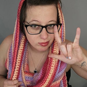 May include: A woman wearing a multi-colored striped crochet scarf with a heart-shaped pendant necklace. She is wearing glasses and has a nose ring.