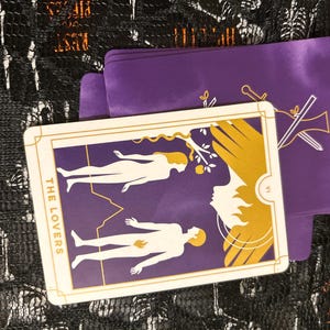 Puede incluir: La carta del tarot Los Enamorados presenta un fondo morado con un contorno blanco de un hombre y una mujer de pie a cada lado de un árbol. La carta está bordeada de oro con las palabras "LOS ENAMORADOS" impresas verticalmente en el lado izquierdo.