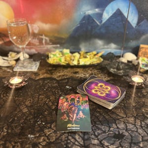 Può includere: Un tovaglia di pizzo nero con due candele accese, un bicchiere d'acqua e due carte dei tarocchi. Una carta mostra uno sfondo viola con un simbolo dorato e l'altra carta mostra un'illustrazione colorata di una donna in un giardino.