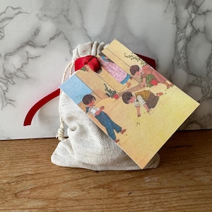 Könnte beinhalten: Ein kleiner weißer Stoffbeutel mit einem roten Band, das um die Oberseite gebunden ist. Der Beutel ist an einer Karte mit einer Vintage-Illustration von Kindern befestigt, die in einem Innenhof spielen.