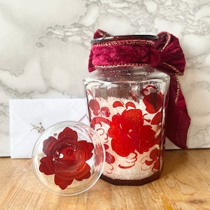 Puede incluir: Un tarro de vidrio transparente con un diseño floral rojo y una cinta de terciopelo rojo atada alrededor de la tapa. El tarro está lleno de cristales blancos y tiene una tapa floral roja a juego.