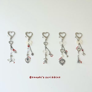 Puede incluir: Cinco llaveros de corazón plateados con diferentes dijes. Los dijes incluyen un lazo, una rosa, un osito de peluche, una llave y la Torre Eiffel. Cada dije está unido a una cadena plateada con una cuenta en forma de corazón.