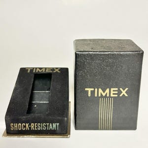 Puede incluir: Caja y expositor de reloj negros con letras doradas. La caja y el expositor llevan la etiqueta "TIMEX". El expositor lleva la etiqueta "SHOCK-RESISTANT". La caja tiene rayas verticales doradas.