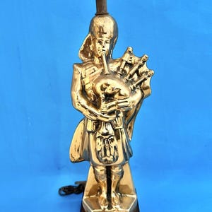 May include: A gold-colored lamp base shaped like a bagpiper playing a set of bagpipes. The base is on a wooden platform with the text "Seagram's 100 Pipers Scotch".