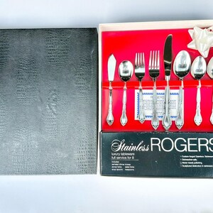 Puede incluir: Un juego de cubiertos de acero inoxidable para ocho personas. El juego incluye cuchillos de mantequilla, cucharas, tenedores y una cuchara de servir. La caja es negra con una apariencia texturizada de cuero sintético. Los cubiertos están dispuestos sobre un fondo rojo.