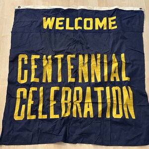 Op de afbeelding: Een blauwe stoffen banner met de tekst "WELCOME CENTENNIAL CELEBRATION" in gele letters.