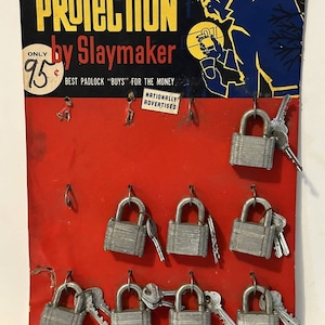 Puede incluir: Una tarjeta de exhibición de cartón roja y negra con el texto "Protection by Slaymaker" y una ilustración de dibujos animados de un hombre sosteniendo un candado. La tarjeta tiene nueve candados plateados con llaves adjuntas, cada uno colgando de un gancho.