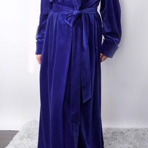 May include: A long, royal blue velvet robe with a matching tie belt. The robe has long sleeves with folded cuffs and a draped front. The soft fabric creates a luxurious look. The robe is suitable for lounging or as a stylish cover-up.