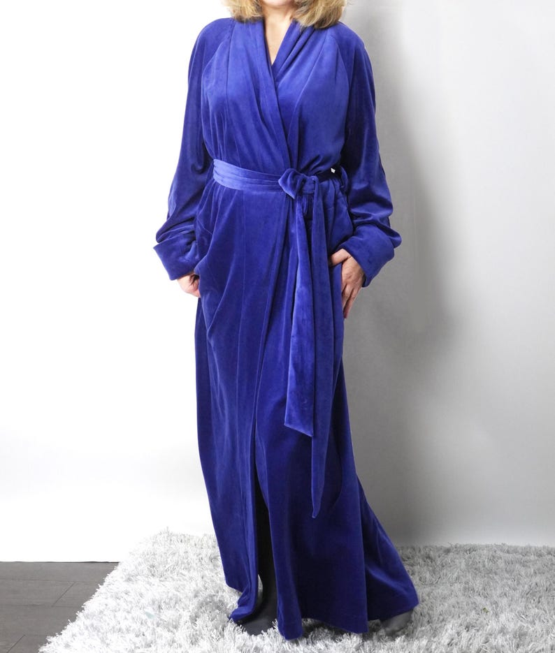 May include: A long, royal blue velvet robe with a shawl collar and a matching waist tie. The robe has long sleeves and a floor-length design. The fabric appears soft and luxurious, suitable for lounging. The robe is shown against a white background.