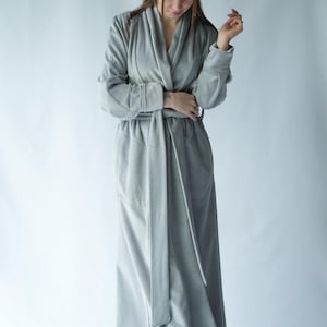 May include: A long, light gray robe with a shawl collar and a matching belt. The robe has long sleeves and a floor-length design. The fabric appears soft and plush, suitable for lounging. The robe is a solid color with a simple, elegant design.