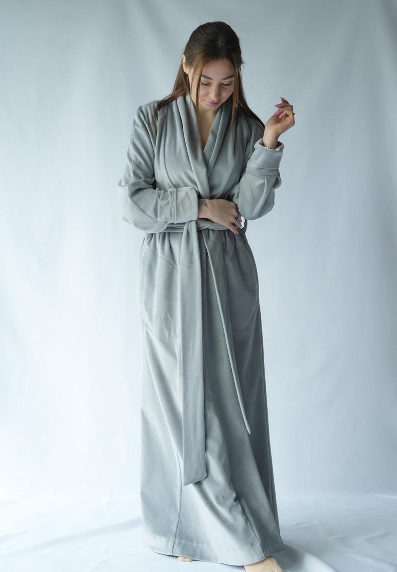 May include: A long, light gray robe with a shawl collar and a matching belt. The robe has long sleeves and a floor-length design. The fabric appears soft and plush, suitable for lounging. The robe is a solid color with a simple, elegant design.